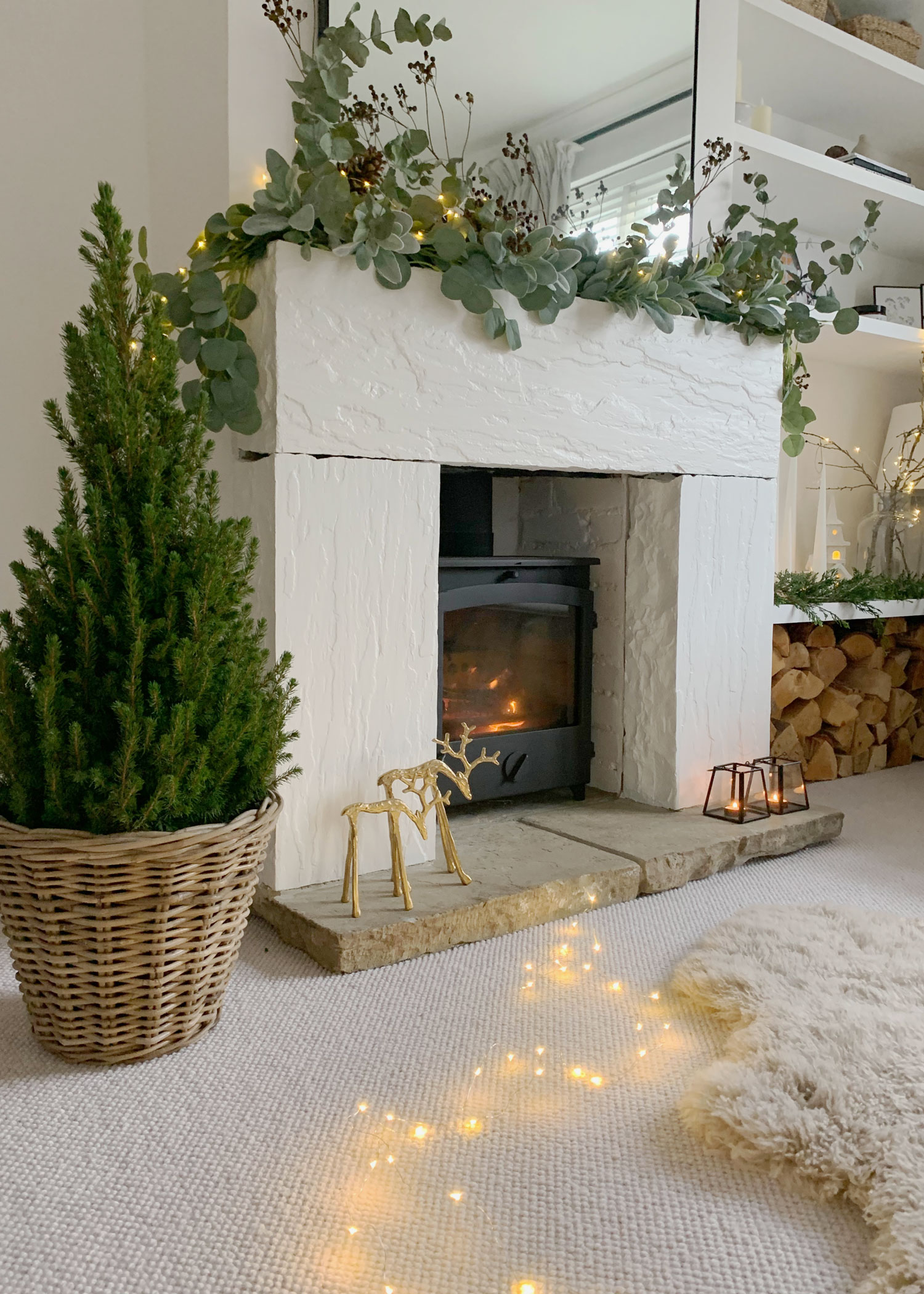 Mini Christmas tree in a wicker basket beside a glowing log burner, styled with twinkling fairy lights and festive eucalyptus garland.