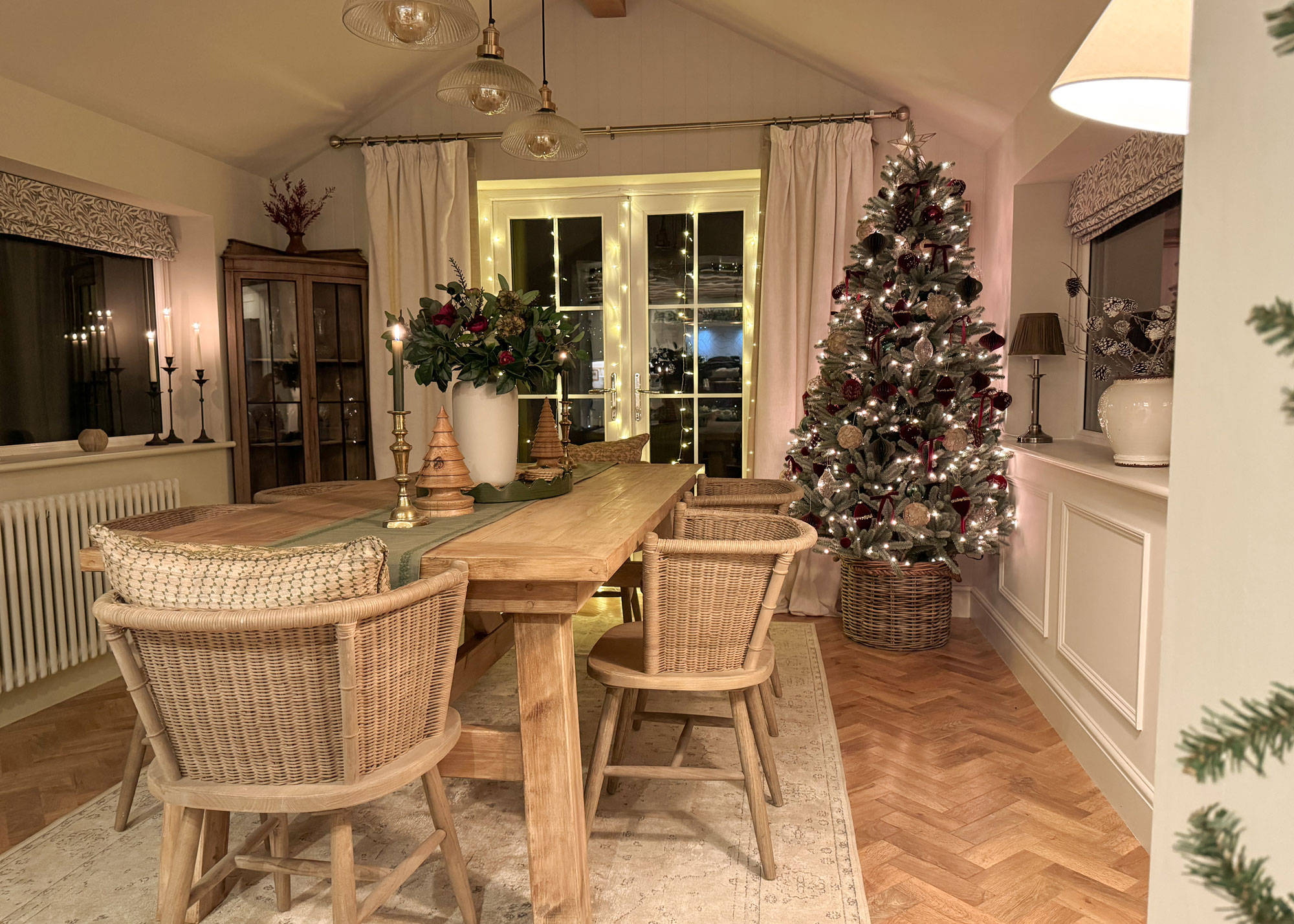 Elegant dining room with a frosted Christmas tree in a wicker tree skirt, surrounded by warm lights and rustic wood furniture.
