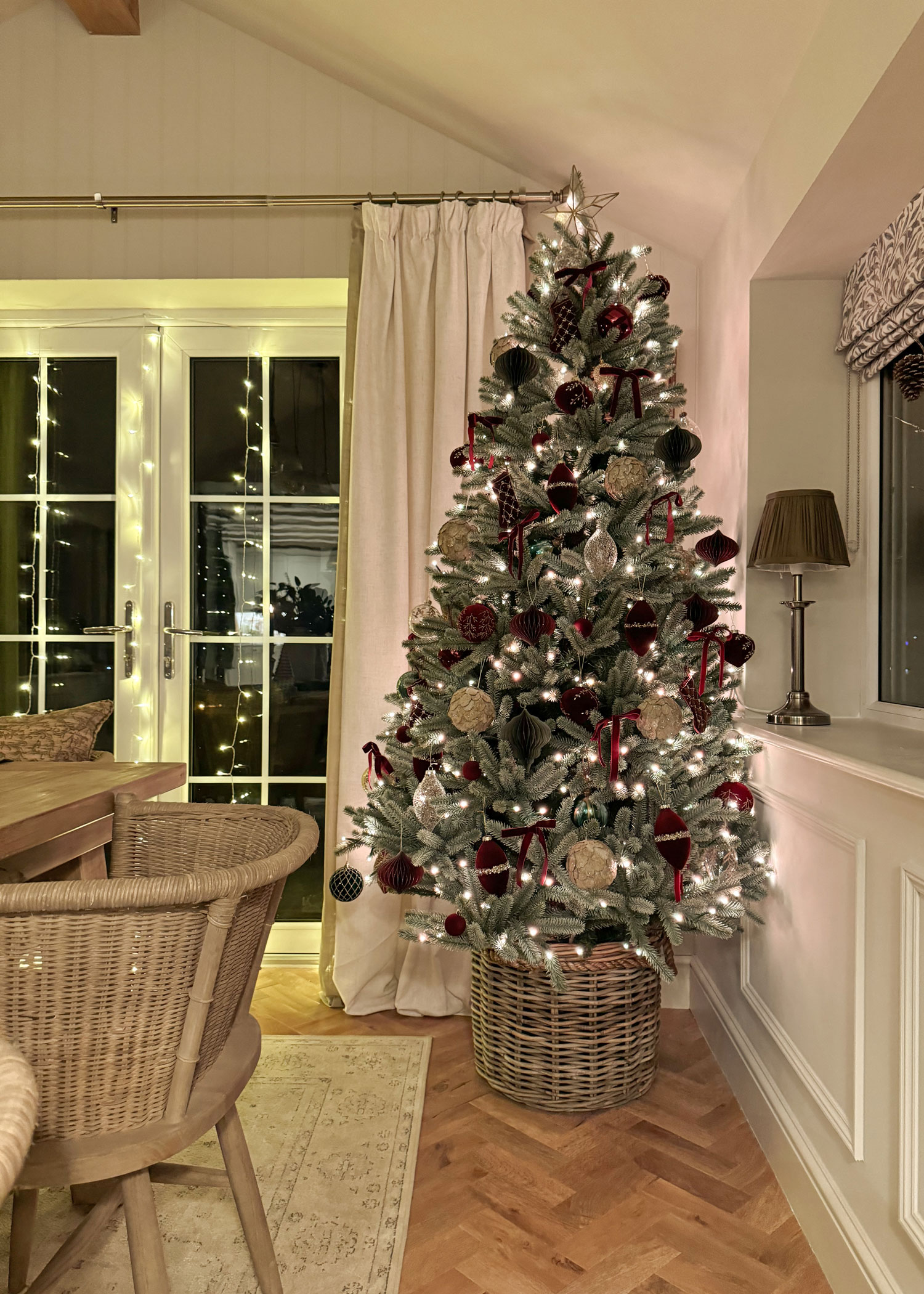 Frosted Christmas tree with red and gold decorations in a wicker tree skirt, glowing warmly in a cosy dining nook.