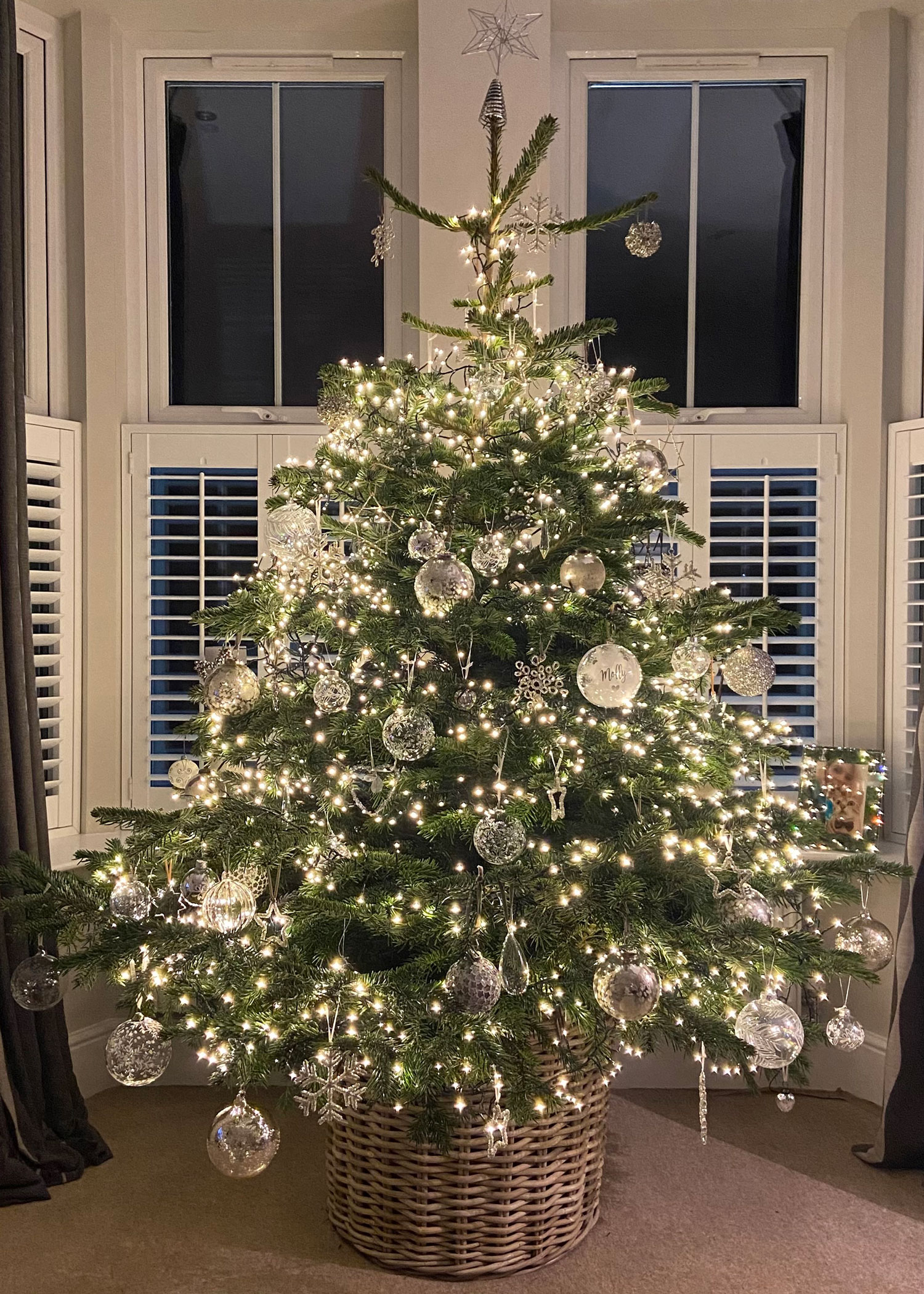 Real Christmas tree with sparkling silver baubles and warm fairy lights, styled in a natural wicker tree skirt at night.