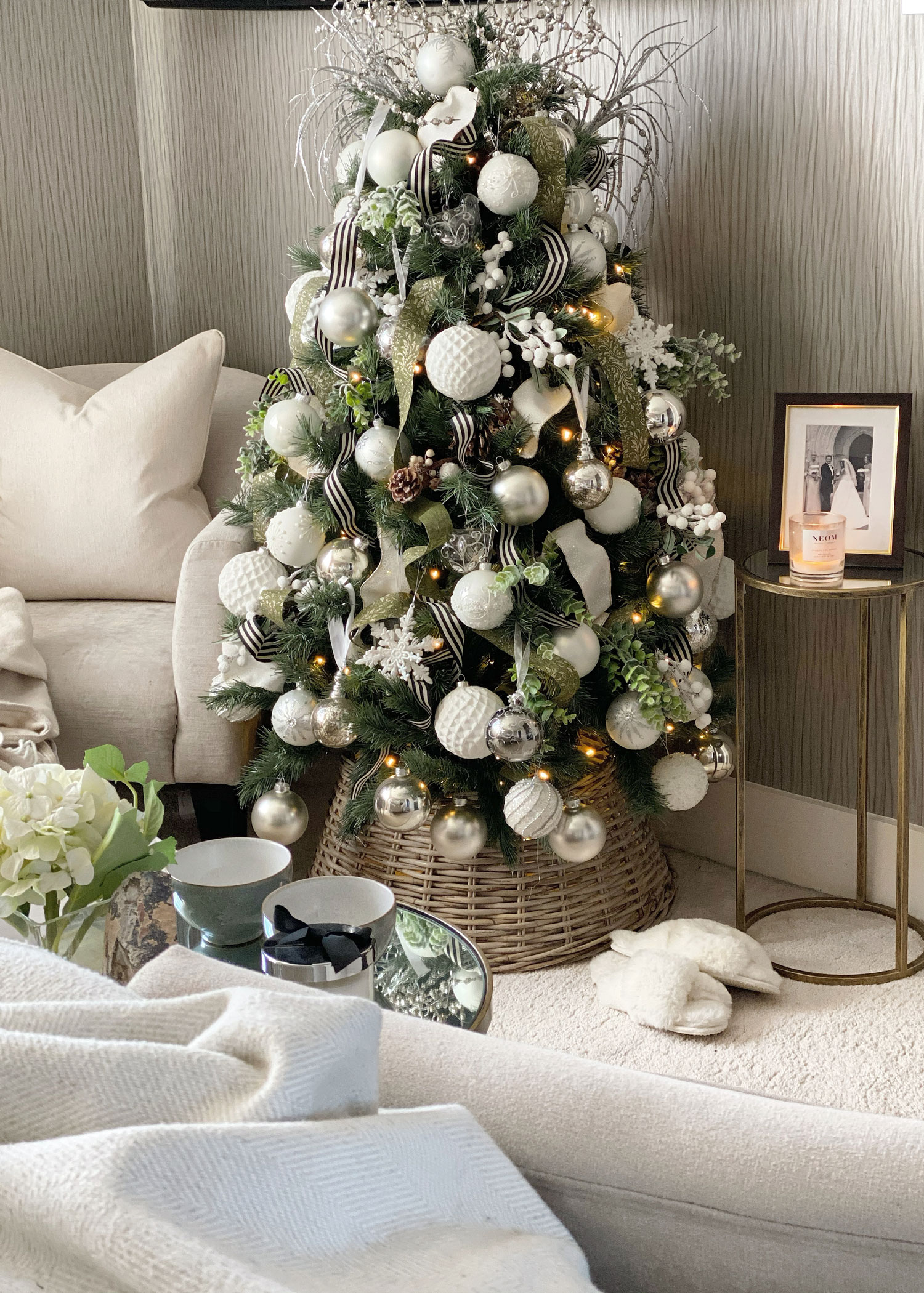 Christmas tree with white and silver baubles, styled in a wicker tree skirt in a calm, neutral-toned living room corner.