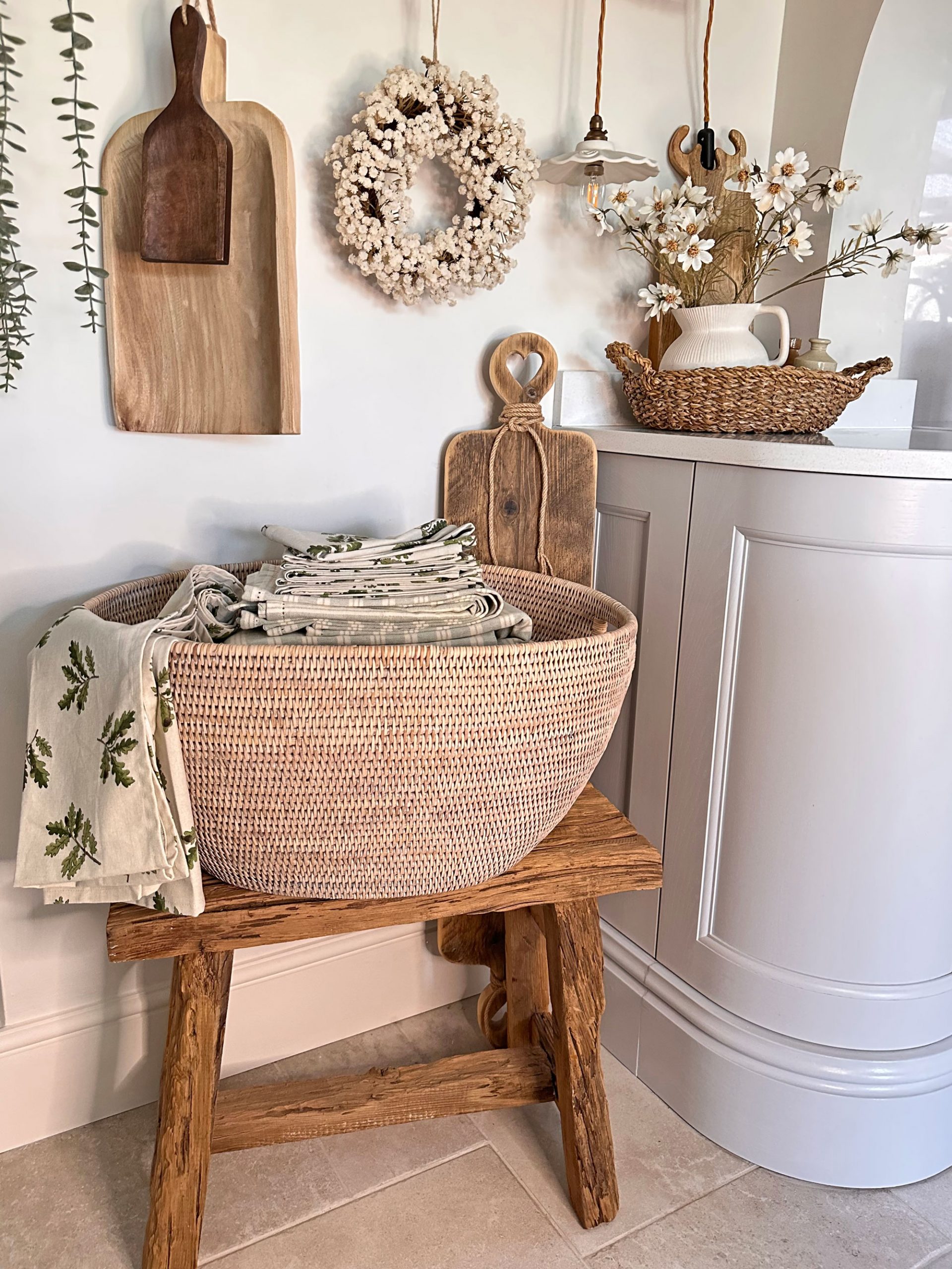 Rustic kitchen corner with a large woven basket filled with folded linens, styled with wooden boards and seasonal floral touches.