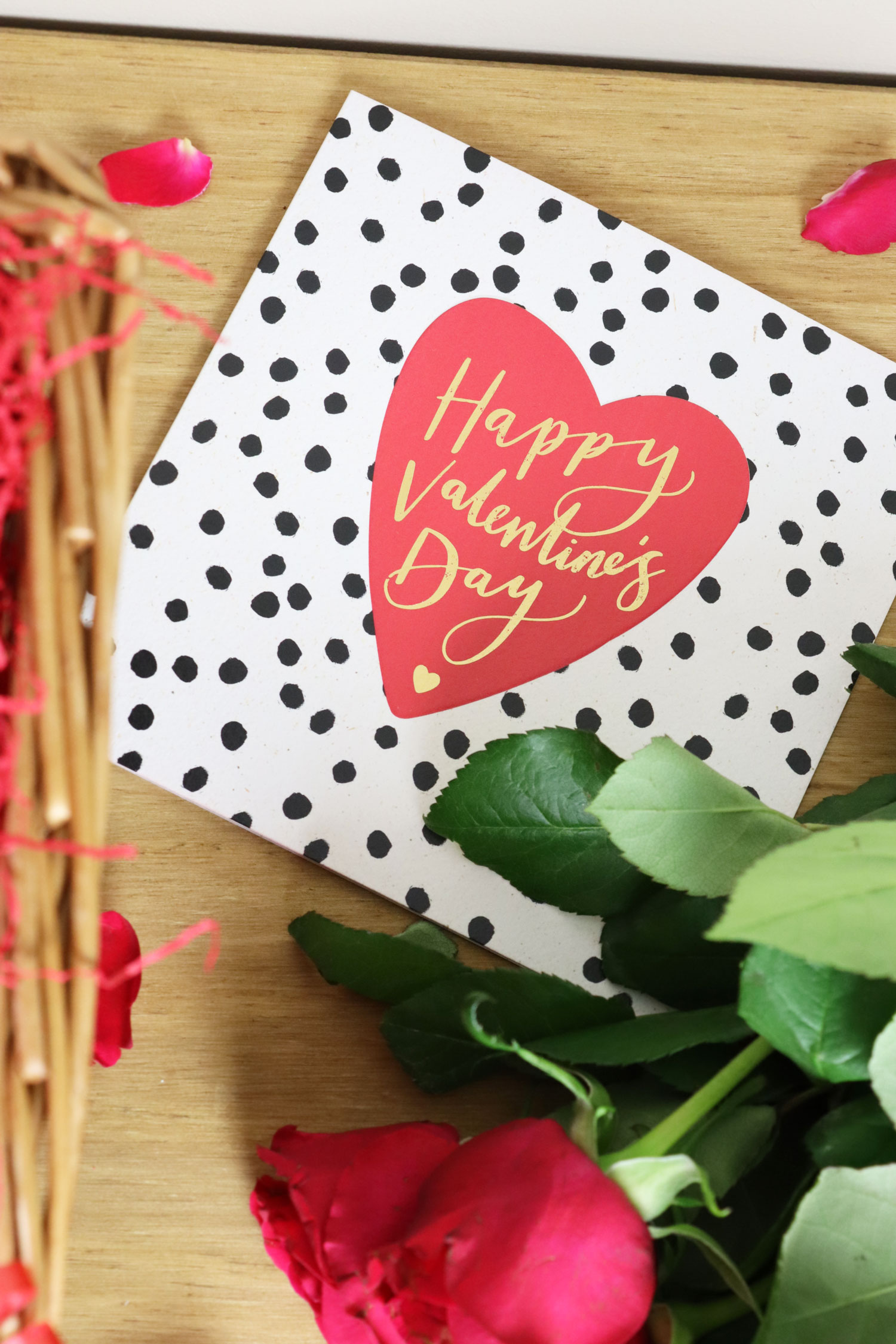 Valentine’s Day card with a red heart design resting beside fresh red roses and rose petals on a wooden surface.