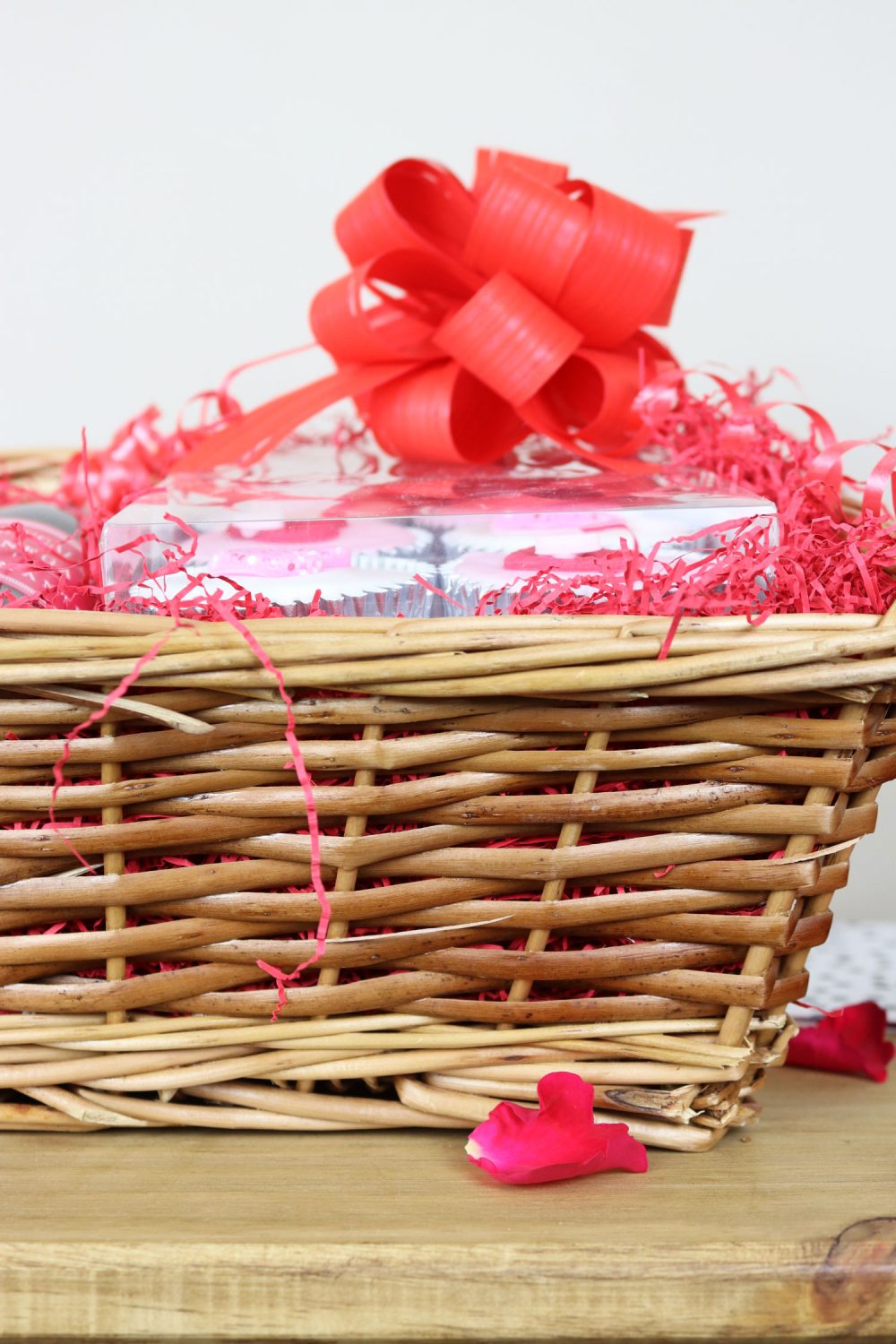 Side view of a wicker Valentine’s hamper basket filled with treats, finished with a large red bow and pink shredded paper.