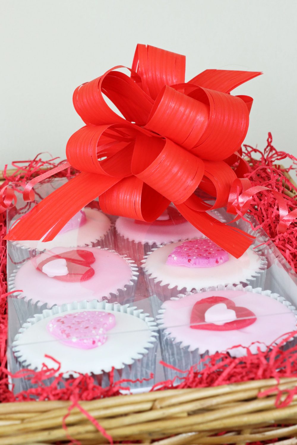 Close-up of pink Valentine’s cupcakes topped with hearts, inside a wicker hamper tied with a bright red ribbon.