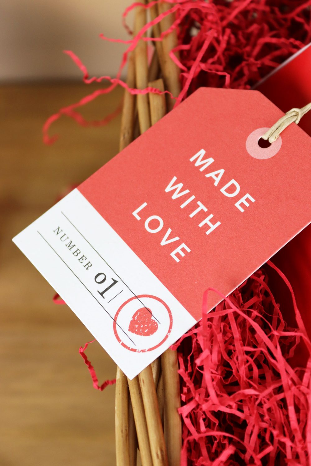Red “Made With Love” gift tag tied to a wicker basket, nestled in red shredded paper for a thoughtful Valentine’s touch.