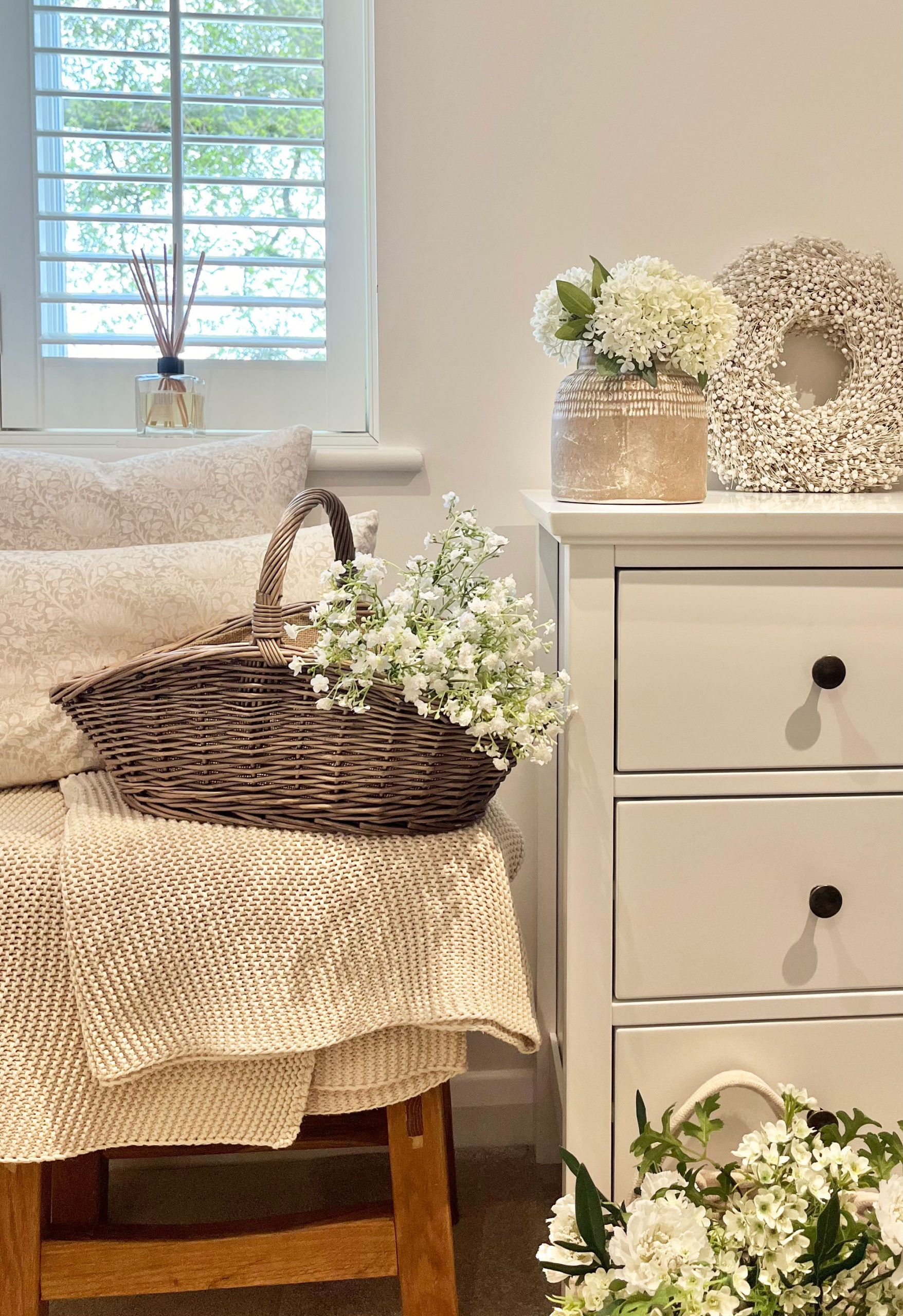 Wicker flower trug filled with white blooms styled in a cosy, neutral-toned bedroom with soft textures.