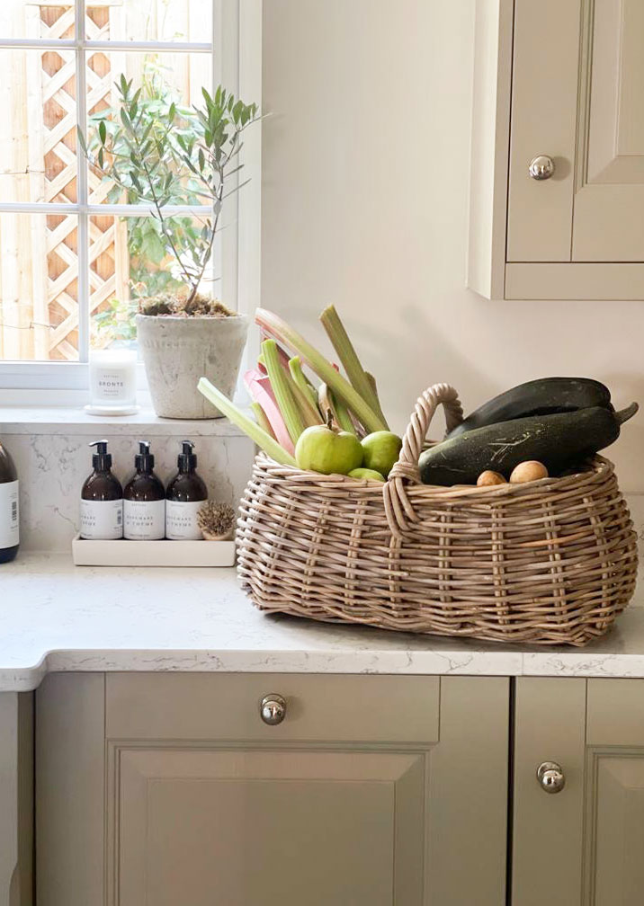 Wicker trug filled with homegrown courgettes, rhubarb and apples on a marble kitchen countertop.