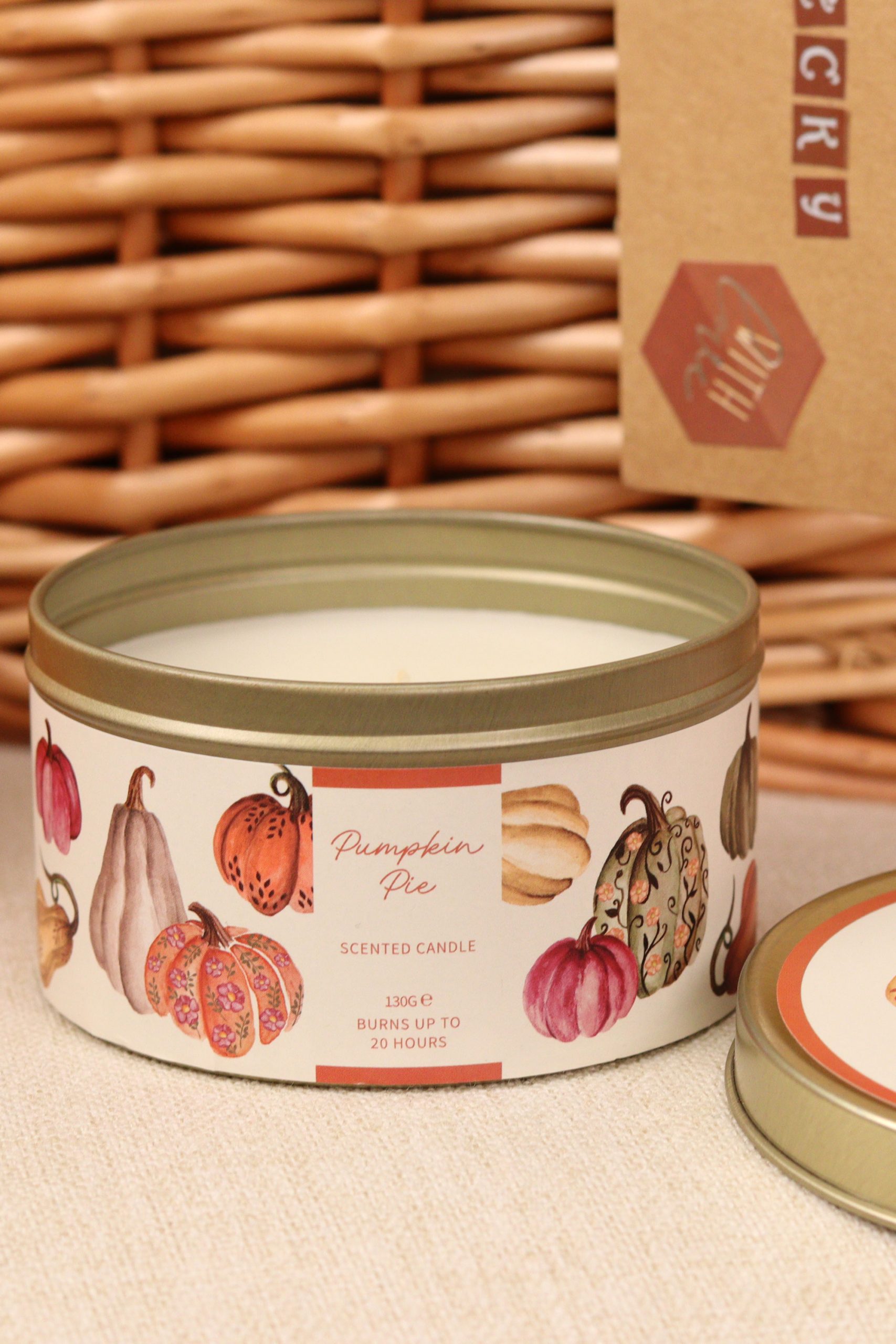 Pumpkin Pie scented candle in a decorative tin with autumn gourd illustrations, placed in front of a wicker basket.