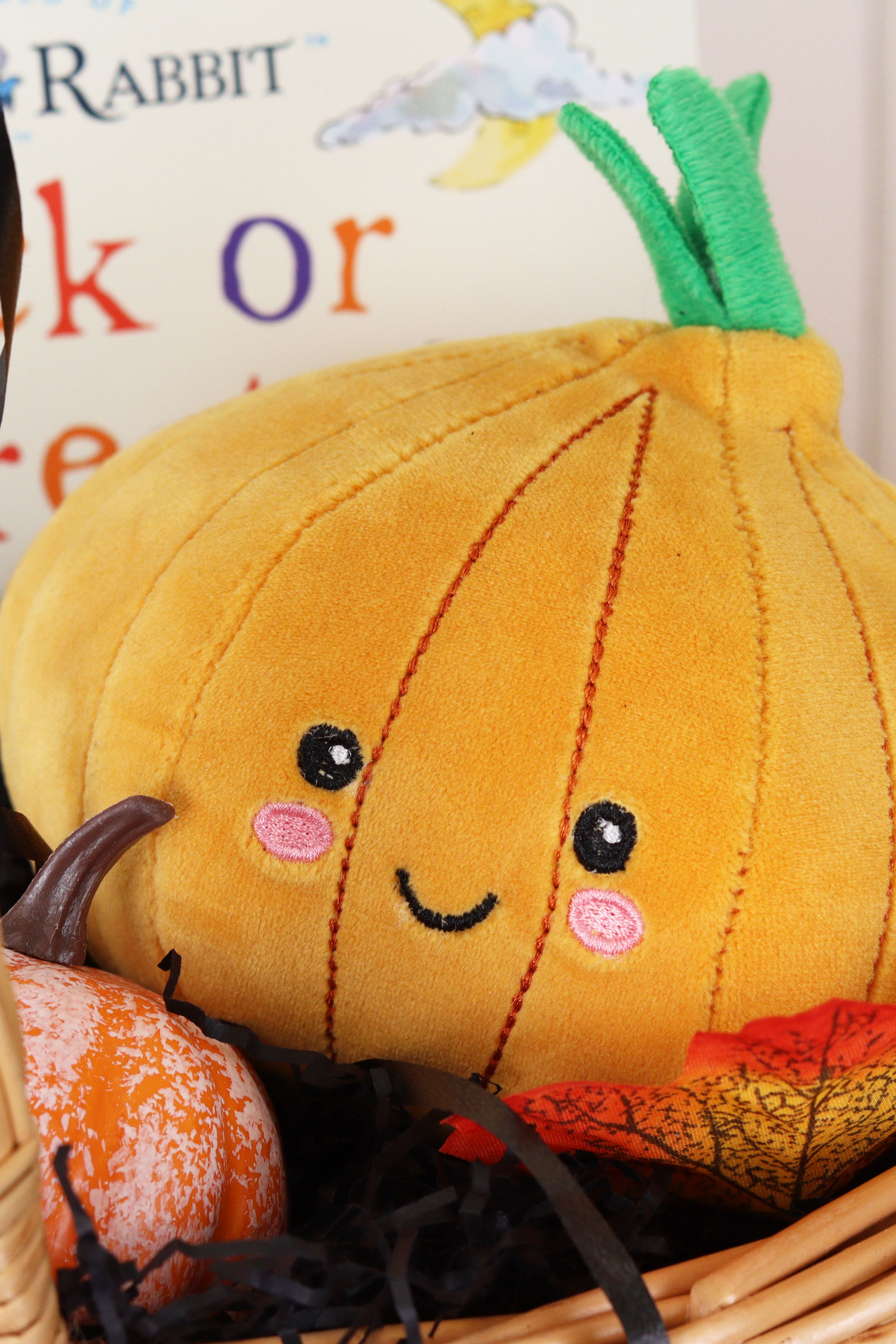 Close-up of a plush pumpkin toy with a smiling face, nestled in a wicker basket with mini pumpkins and autumn leaves.