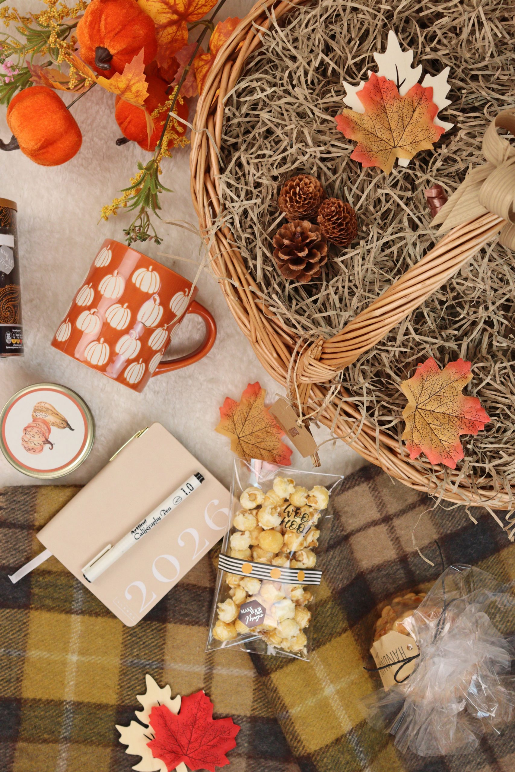 Flat lay of autumn gift basket contents including a pumpkin mug, popcorn, cookies, 2026 diary, candle, and leaves on a cosy blanket