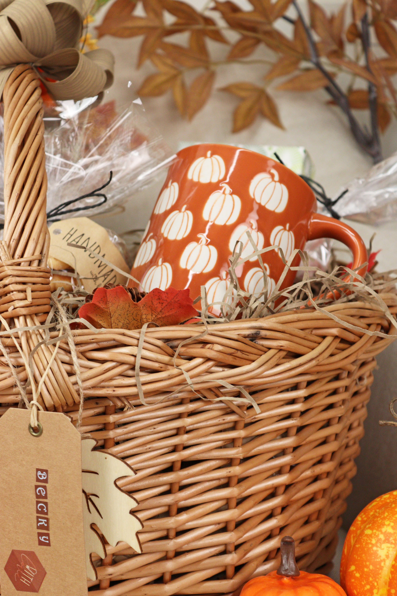 Close-up of a wicker gift basket with an orange pumpkin patterned mug nestled among shredded paper, autumn leaves and seasonal treats