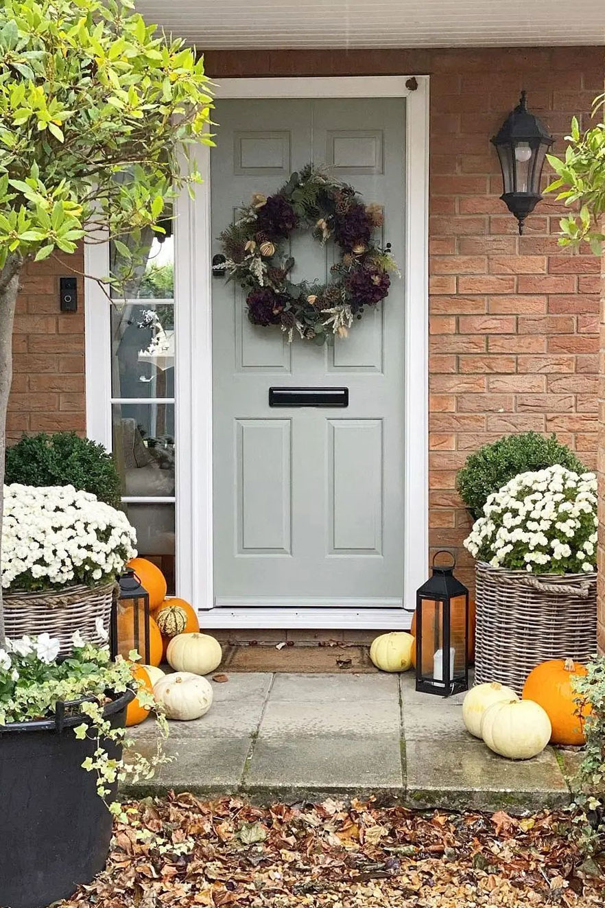 Cosy autumn porch with white chrysanthemums in large wicker planters, surrounded by pumpkins and a sage green front door.