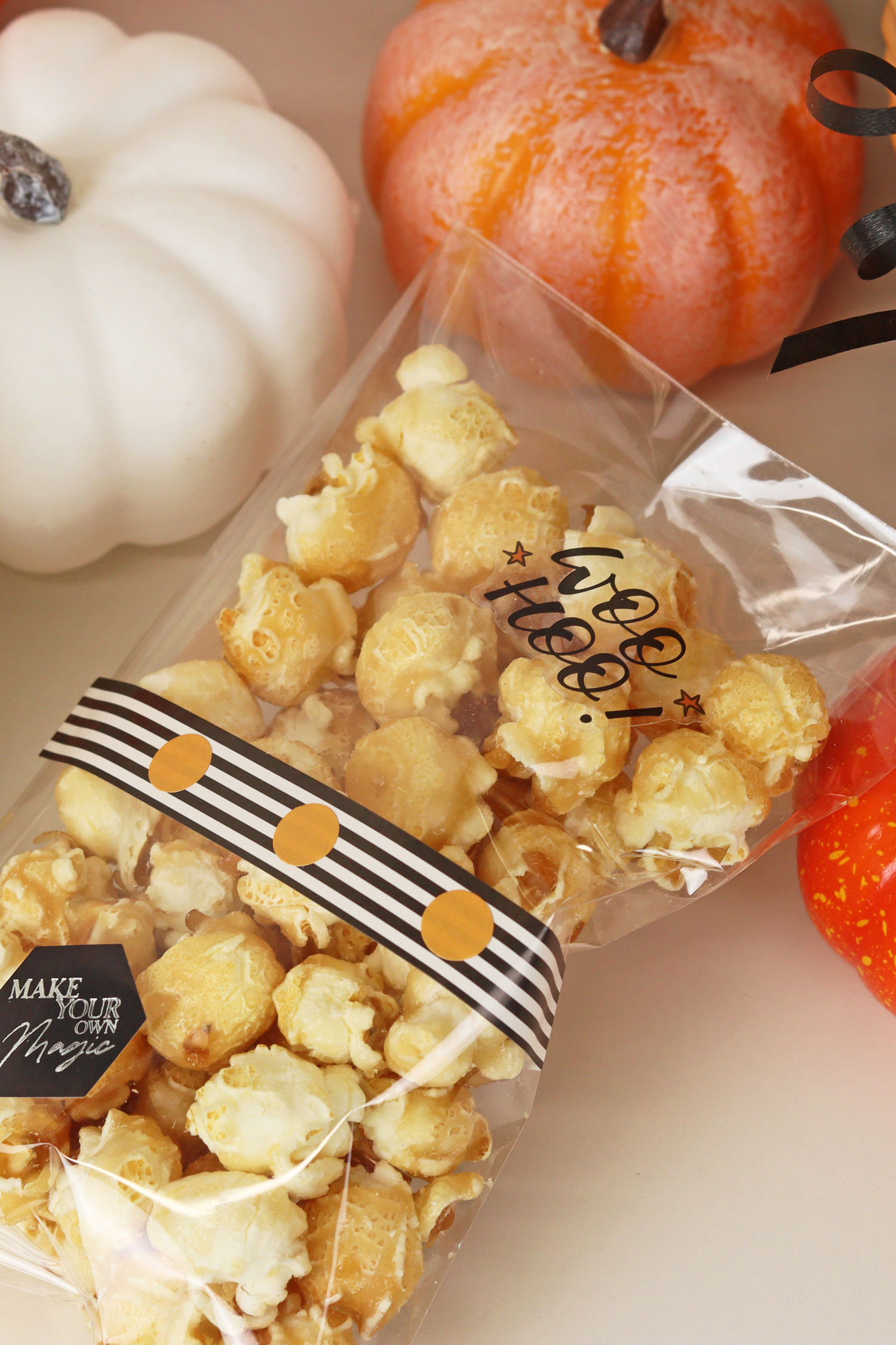 Bag of caramel popcorn with a 'Woo Hoo!' label, decorated with striped ribbon, surrounded by mini pumpkins.