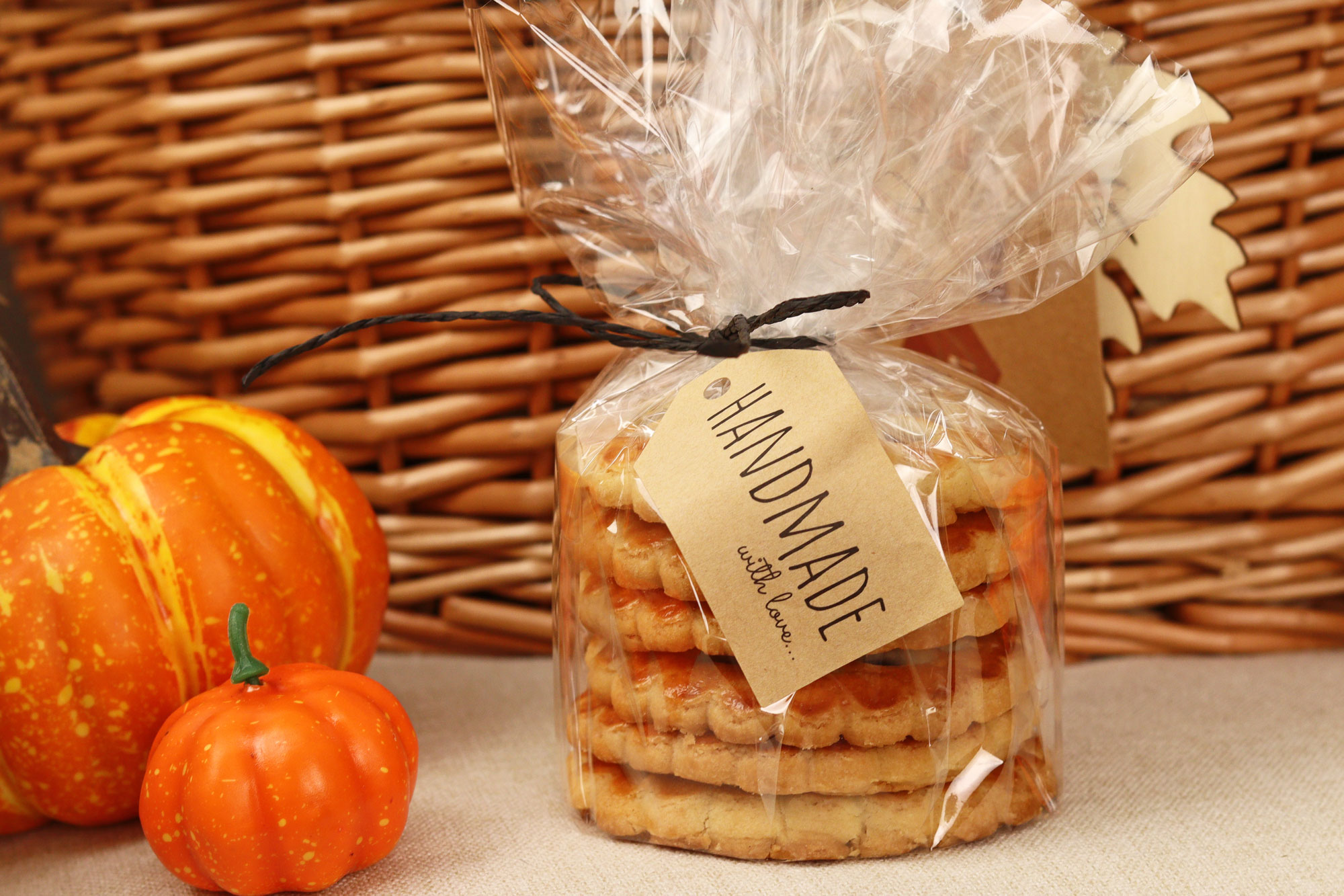 Stack of handmade apple pies wrapped in clear cellophane with a kraft tag, placed beside mini decorative pumpkins and a wicker basket