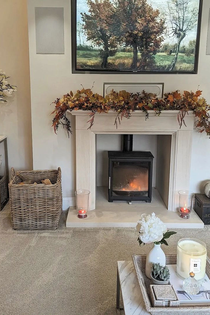 Elegant cream fireplace with a black wood burner, autumn garland and a tall wicker log basket filled with firewood.