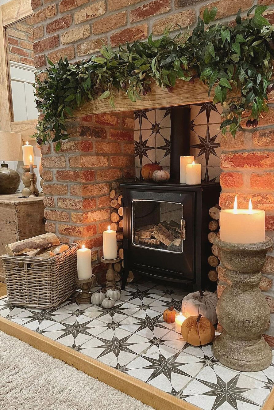 Rustic brick fireplace with a black wood burner, surrounded by candles, pumpkins, and a wicker log basket filled with firewood.