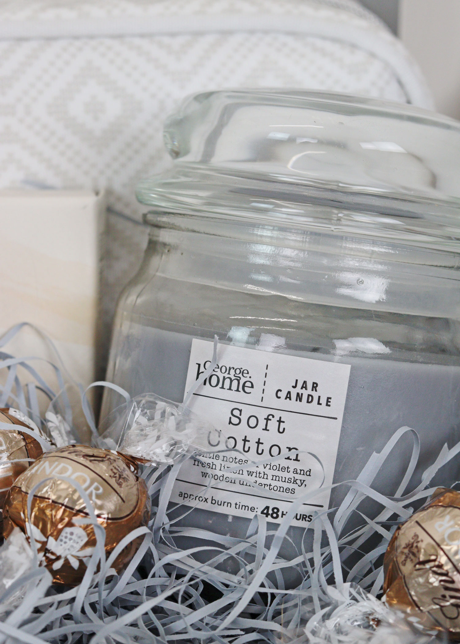 Soft Cotton jar candle nestled in shredded paper inside a burr basket, styled with Lindor chocolates and cosy textures.