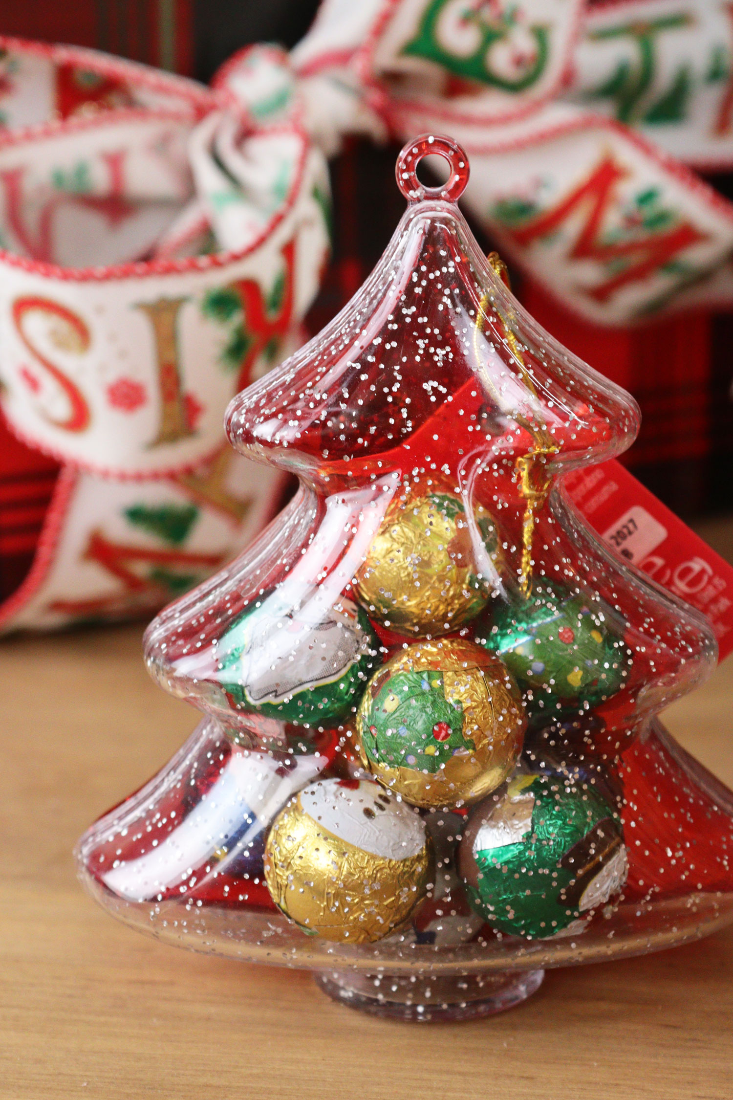 Clear Christmas tree ornament filled with red, gold and green foil-wrapped chocolate truffles on a wooden table.