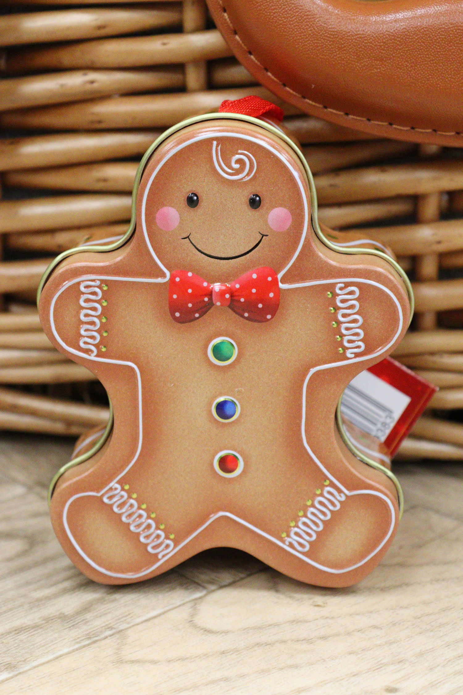 Gingerbread man gift tin with red bowtie and candy buttons, placed in front of a wicker Christmas hamper.