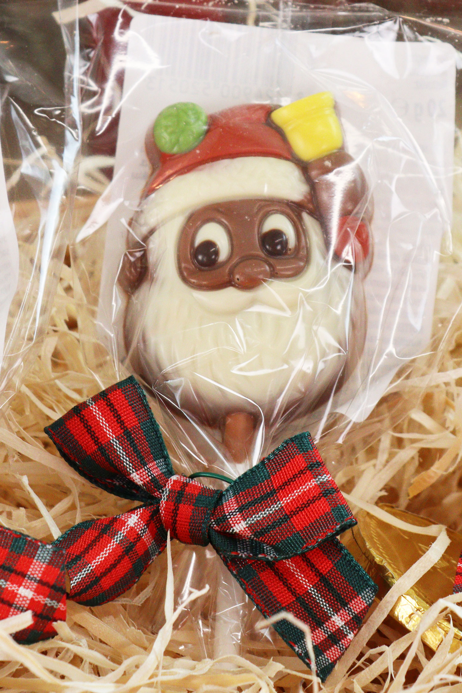 Close-up of festive Father Christmas chocolate lolly with tartan ribbon, nestled in shredded paper gift hamper filling.