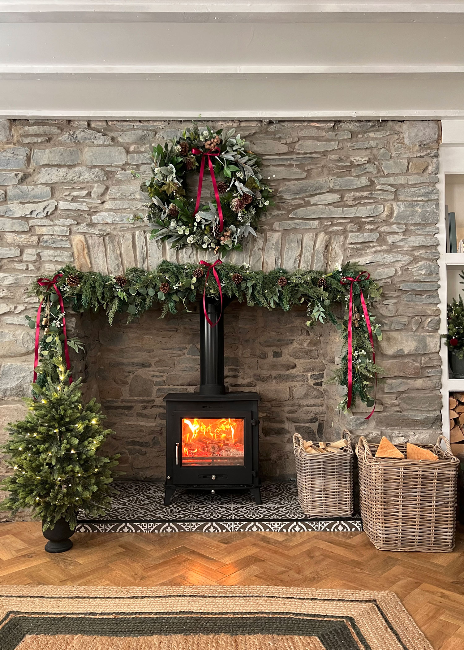Stone fireplace decorated for Christmas with a glowing wood burner, festive greenery, and two wicker log baskets filled with firewood.