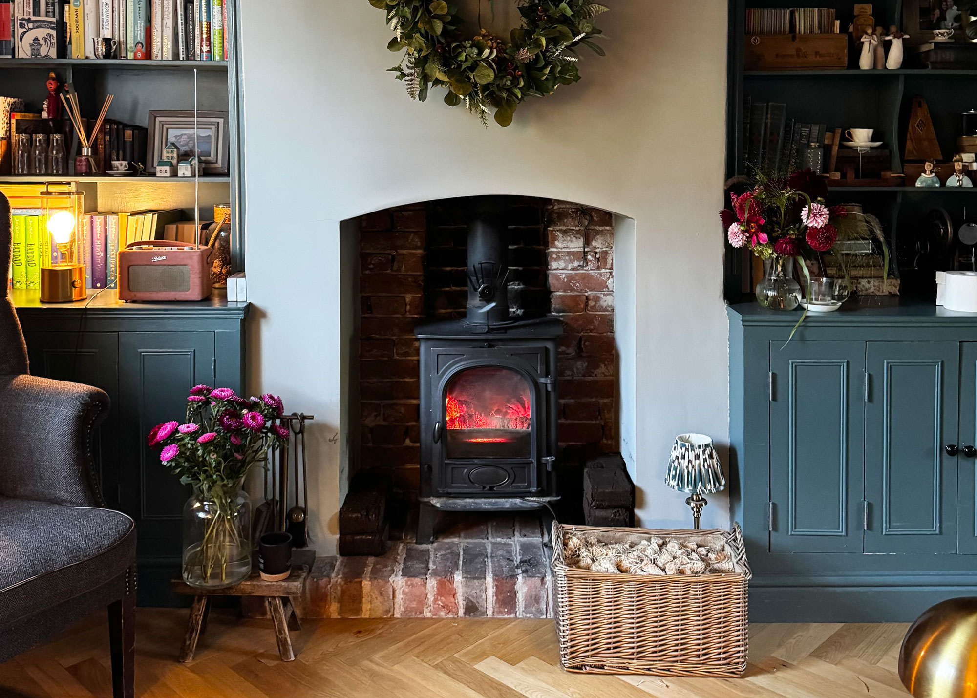 Characterful living room with a red-brick fireplace, glowing wood burner, and wicker log basket filled with kindling beside built-in shelves.
