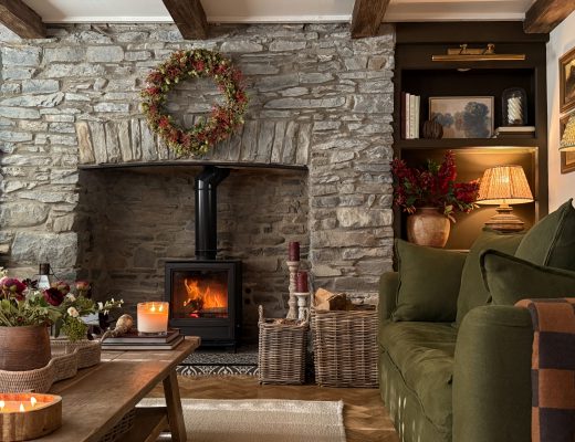 Cosy stone fireplace with a glowing wood burner, flanked by a large wicker log basket filled with firewood.