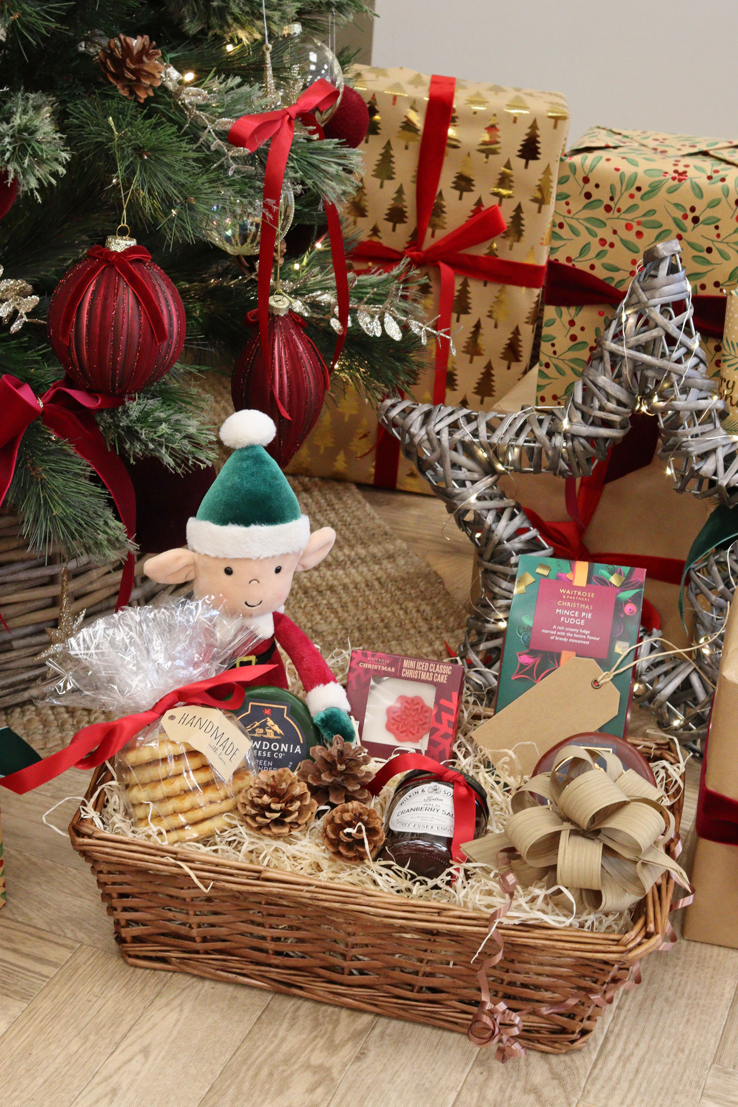 Small festive wicker hamper with cheese, crackers, mulled wine mix and ribbon under a decorated Christmas tree.