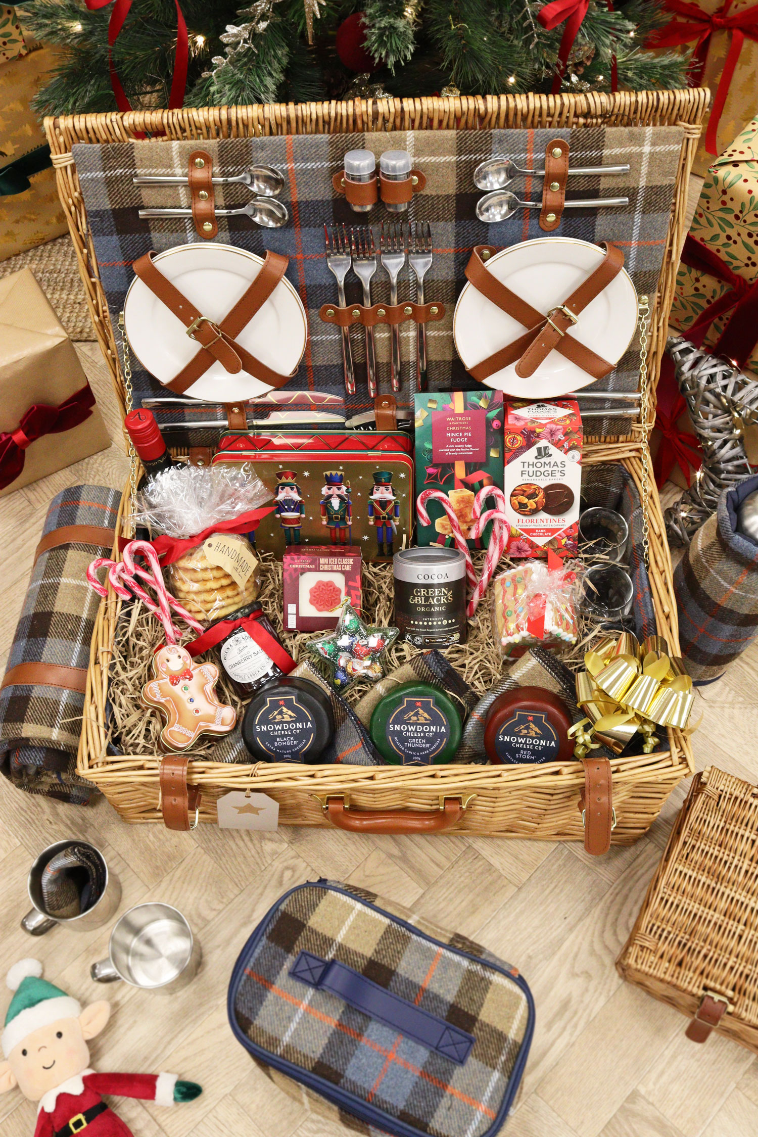 Large Christmas food hamper open to reveal luxury cheeses, treats and plates, styled beside a tartan blanket.