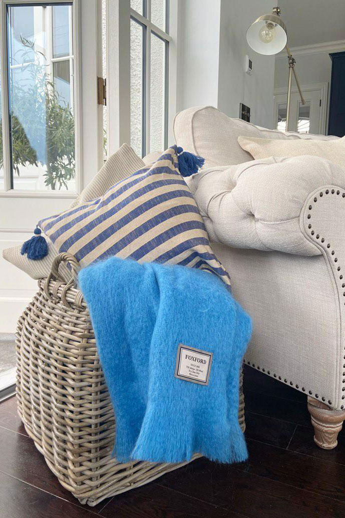 Grey & buff rattan blanket basket styled beside a cream sofa, holding bold striped cushions and a bright blue throw.