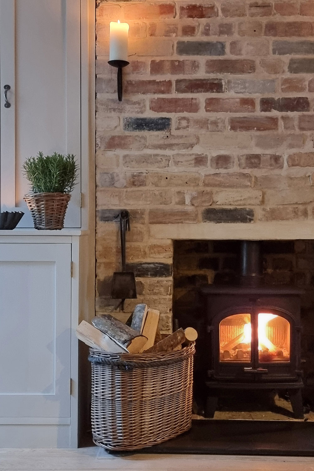 Traditional brick fireplace with a curved wicker log basket filled with chopped firewood, styled for a warm rustic feel.