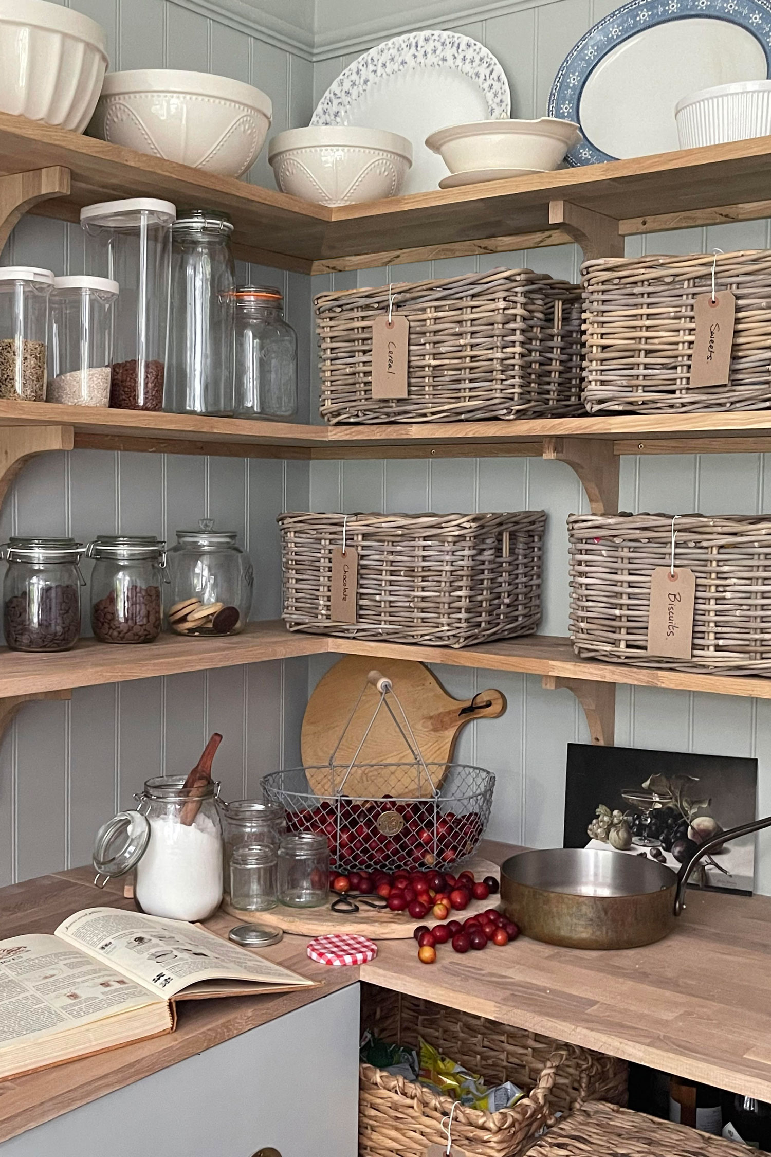 Open wooden kitchen shelves neatly arranged with glass jars and rectangular rattan storage baskets for a warm, practical pantry feel.