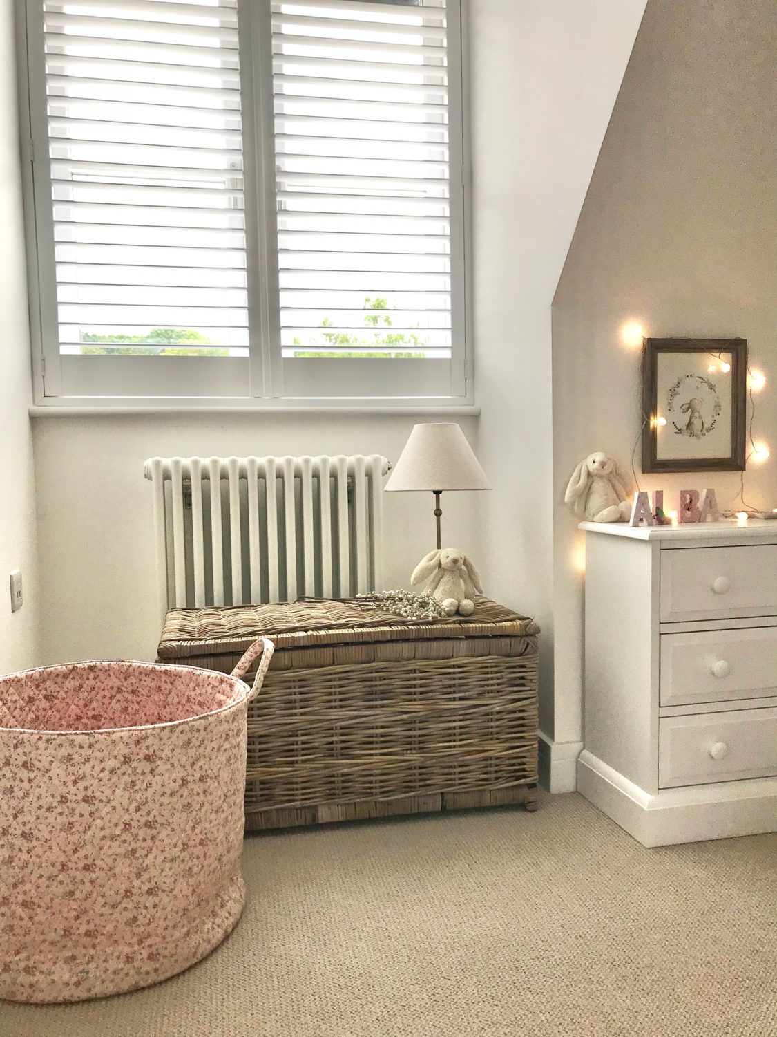 Softly lit nursery with a rattan storage trunk used as a toy basket, paired with a pink fabric laundry bin and warm neutrals.