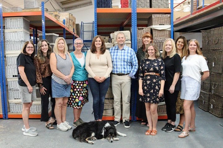 The Basket Company team standing together in the warehouse, surrounded by neatly stacked wicker baskets and storage solutions.