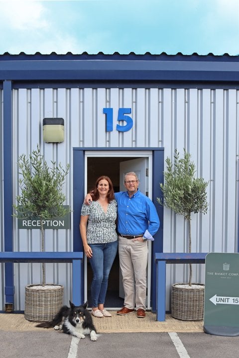 The Basket Company directors, Catherine and Ian, standing outside The Basket Company reception entrance with olive trees in wicker planters. They are joined by their dog Alfie. 